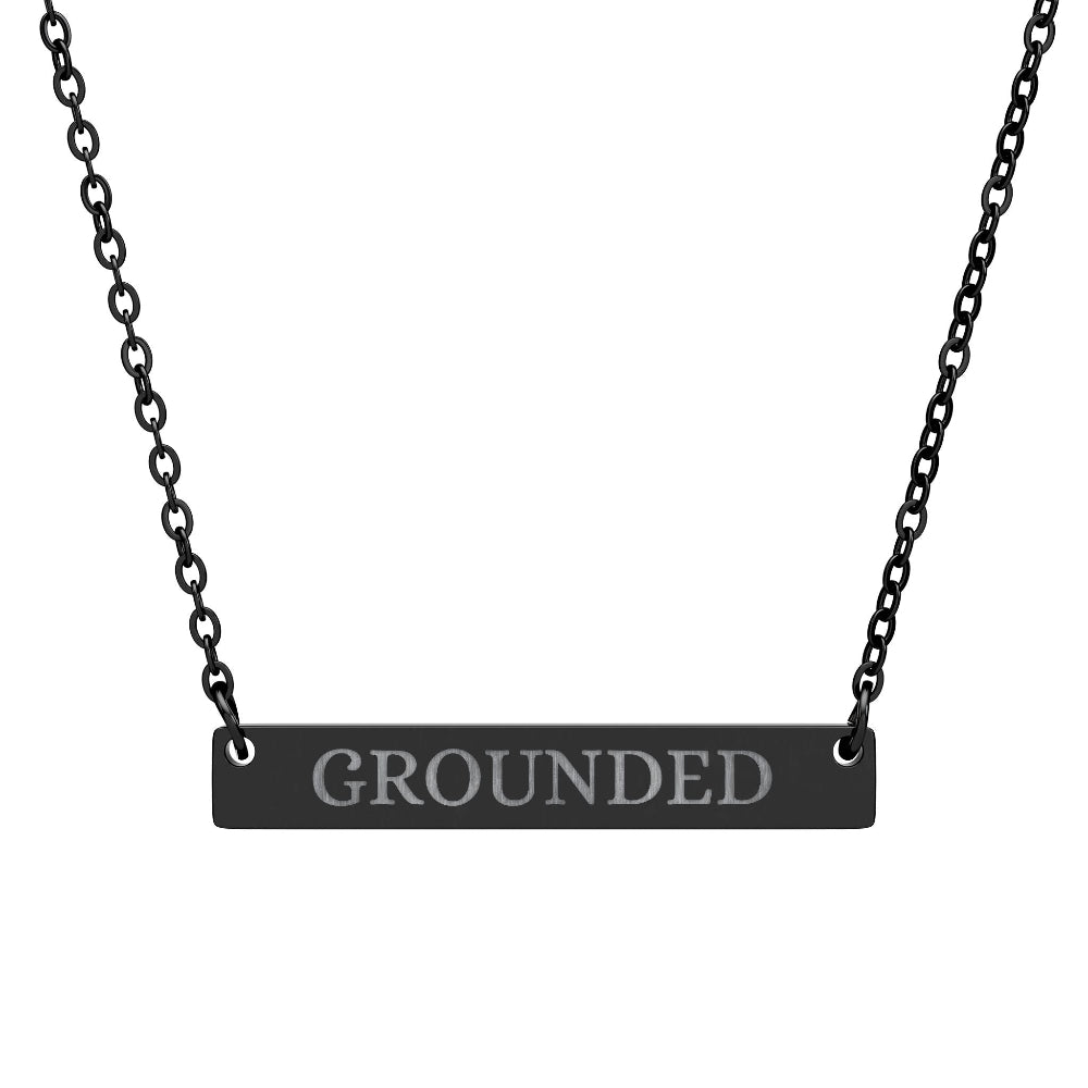 Black bar necklace with engraved uppercase text "GROUNDED" on a matte black pendant, suspended on a delicate black chain. Minimalist motivational affirmation jewelry for grounding, stability, and root chakra energy.