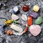 Chakra healing crystal set on natural rock surface – rose quartz heart, tumbled gemstones (sodalite, carnelian, citrine, green aventurine, amethyst, clear quartz), gold-wrapped quartz point pendant on black cord, and rainbow chakra chip bracelet, spiritual wellness collection