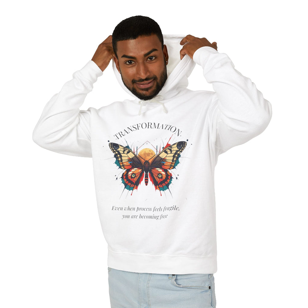 Young man modeling white hoodie with colorful abstract butterfly graphic, "TRANSFORMATION" arched above, and inspirational quote "Even when process feels fragile, you are becoming free" below, pulling up hood