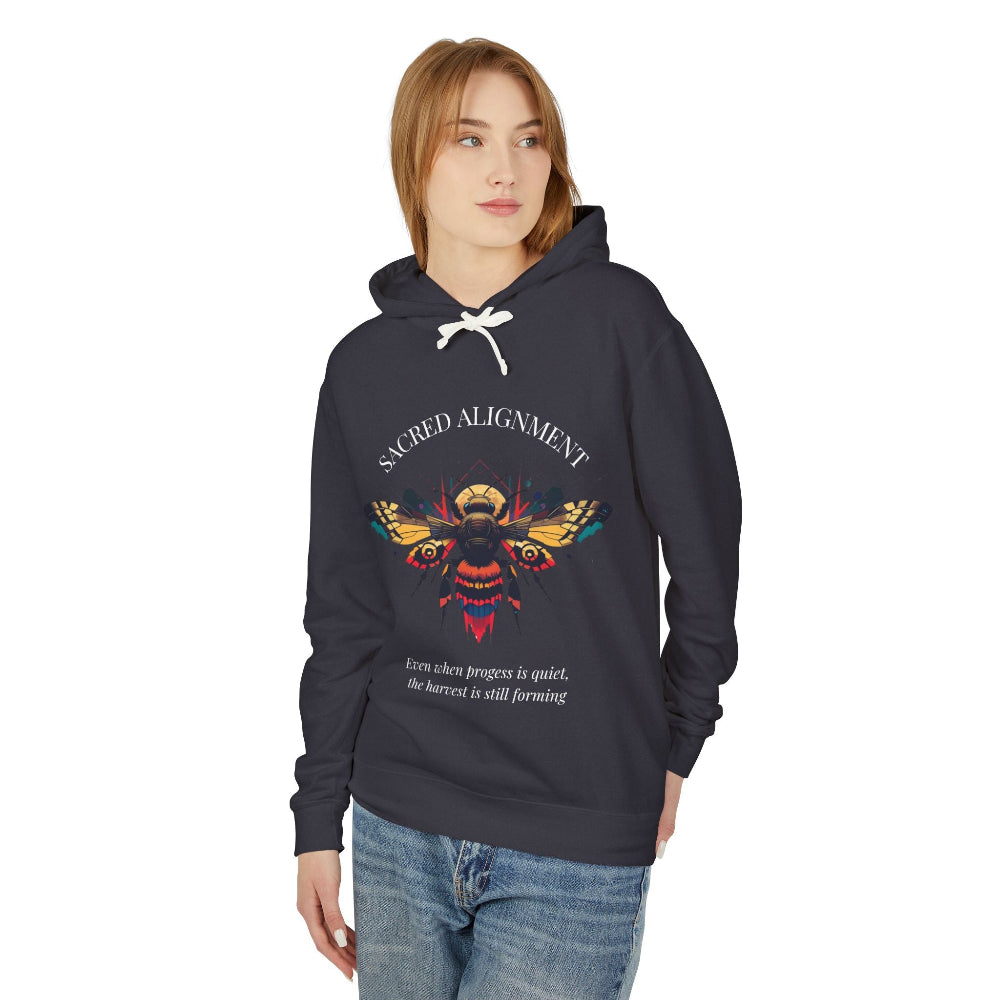 Woman wearing a black hoodie featuring a vibrant bee graphic with the text "SACRED ALIGNMENT" above and the quote "Even when progress is quiet, the harvest is still forming" below. Casual streetwear style with blue jeans.
