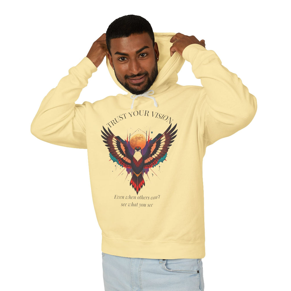 Young man modeling light yellow hoodie with colorful abstract eagle/phoenix graphic, "TRUST YOUR VISION" arched above, and inspirational quote "Even when others can't see what you see" below, pulling up hood