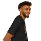 Black unisex cotton t-shirt in side profile on smiling male model with curly hair and beard, featuring bold "Primal" graphic on chest: stylized intense bearded man portrait with short styled hair, vibrant multicolored geometric face in red, blue, teal, orange and yellow tones, dynamic paint splatters bursting outward. Primal masculine energy motivational crewneck tee.