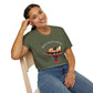 Woman wearing an olive green t-shirt with a vibrant hummingbird graphic, "MOVE WITH GRACE" text above, and the quote "Even when the world accelerates, Your center remains steady" below. Casual motivational apparel paired with blue jeans.
