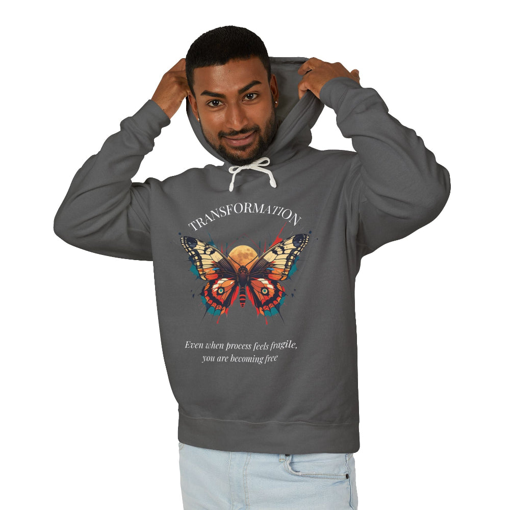 Young man modeling charcoal gray hoodie with colorful abstract butterfly graphic, "TRANSFORMATION" arched above, and inspirational quote "Even when process feels fragile, you are becoming free" below, pulling up hood