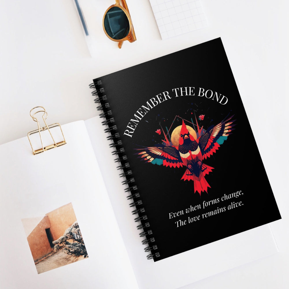 Spiral notebook with 'Remember The Bond' inspirational bird cover design and quote 'Even when forms change, The love remains alive' styled on a white desk with accessories.