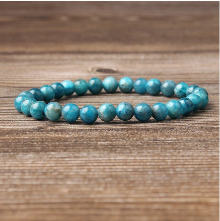 Blue apatite gemstone bracelet on wooden background: polished round beads in vibrant turquoise-blue with natural cloudy inclusions, shiny faceted surfaces, and subtle chatoyancy. Natural healing crystal jewelry for throat chakra, communication, and emotional clarity.