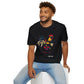 Black unisex cotton t-shirt modeled by smiling man with curly hair and beard, seated pose, featuring bold Year of the Rooster zodiac graphic: vibrant abstract rooster portrait with detailed feathers, proud stance, intense eyes, red comb and wattles, geometric accents, and dynamic multicolored paint splatters in gold, red, black, blue, teal, and orange tones exploding outward. Chinese text "鸡年" (Year of the Rooster) printed below in white. Zodiac Chinese New Year motivational crewneck tee.