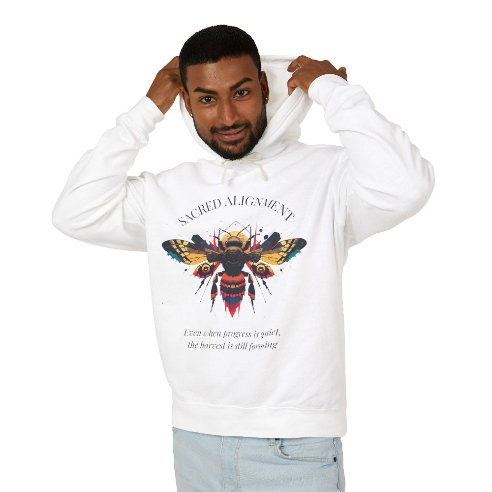 Man wearing a white hoodie featuring a vibrant bee graphic with the text "SACRED ALIGNMENT" above and the quote "Even when progress is quiet, the harvest is still forming" below. Casual streetwear style with light jeans.