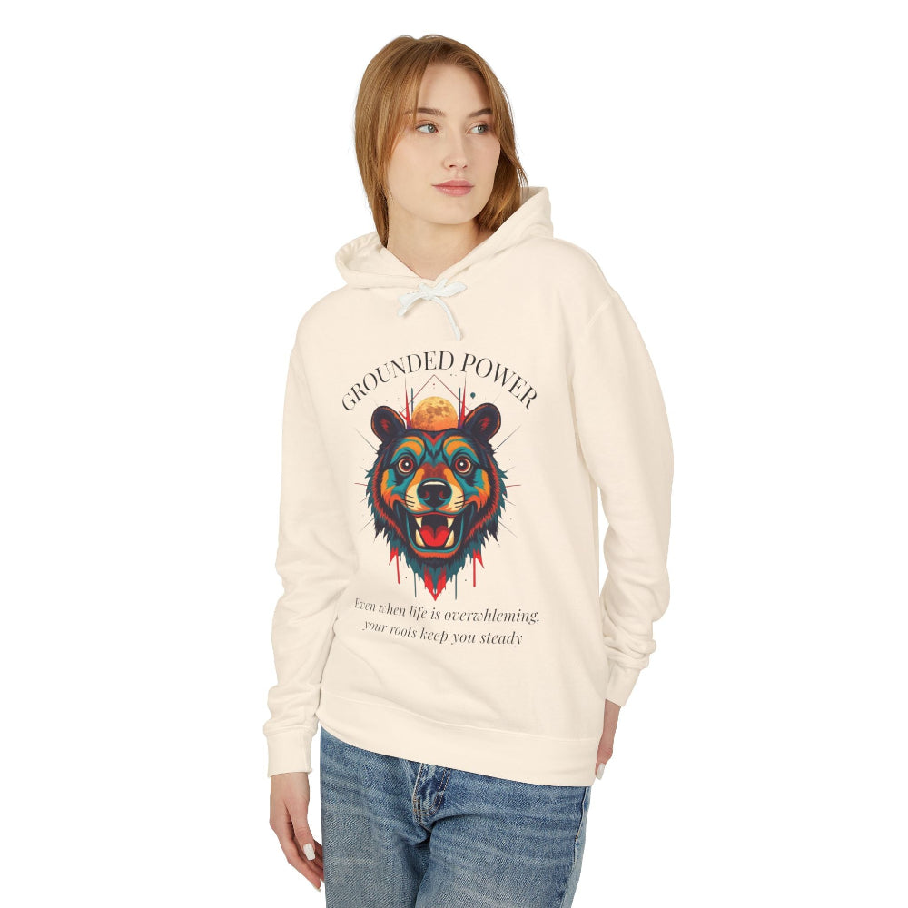 Young woman with long hair modeling cream hoodie featuring vibrant multicolored bear head graphic, roaring expression, fur in blue orange teal splashes, orange moon above, "GROUNDED POWER" arched text, motivational quote about roots and steadiness below