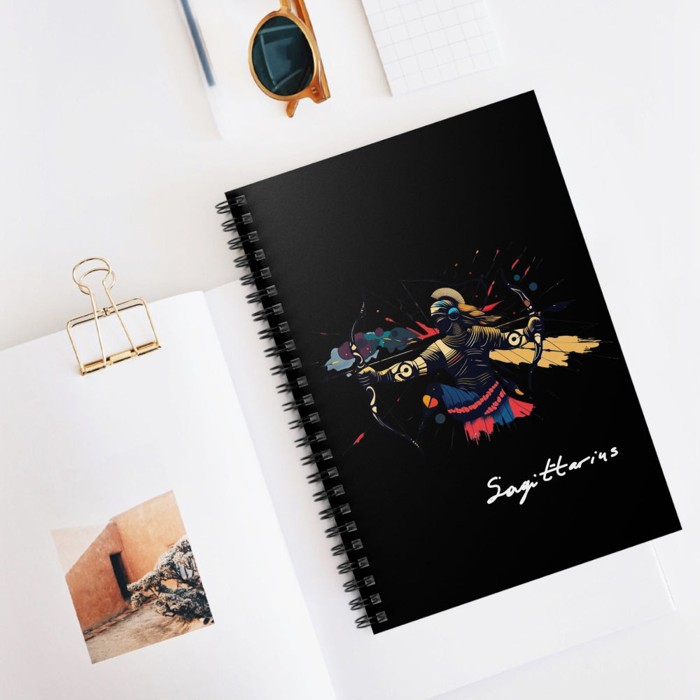 Notebook with a colorful abstract archer drawing a bow in dynamic warrior pose, flowing cape, and paint splatters on a black background, featuring the word "Sagittarius" in elegant white cursive, placed on a minimal white desk with sunglasses and stationery items.