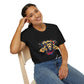 Black unisex cotton t-shirt modeled by smiling woman with braided hair, seated pose, hand on cheek, featuring bold Leo zodiac graphic: vibrant abstract lion head portrait with powerful flowing mane, fierce intense eyes, regal expression, geometric accents, and dynamic multicolored paint splatters in gold, red, navy, teal, and black tones exploding outward. Elegant white cursive "Leo" text below. Astrology zodiac motivational crewneck tee.