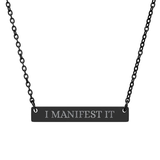Black bar necklace with engraved uppercase text "I MANIFEST IT" on a matte black pendant, suspended on a delicate black chain. Minimalist motivational affirmation jewelry for manifestation and intention setting.