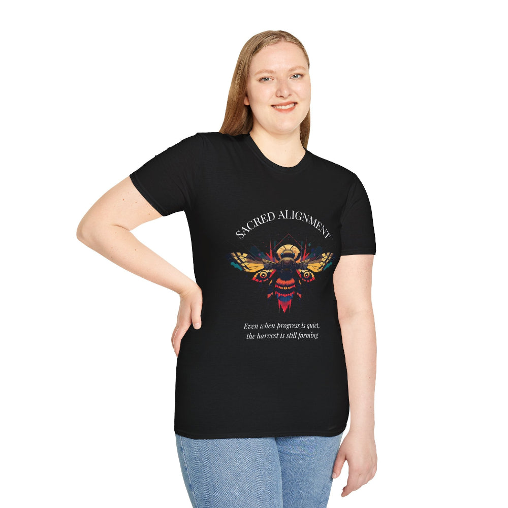 Black plus-size unisex cotton t-shirt modeled by smiling woman with long blonde hair, hand on hip, featuring bold "Sacred Alignment" graphic on chest: vibrant abstract bee with detailed wings and body, colorful geometric accents, and dynamic paint splatters in red, orange, teal, blue, and gold tones around an orange moon. Arched white text "SACRED ALIGNMENT" above and smaller quote "Even when progress is quiet, the harvest is still forming" below. Motivational spiritual empowerment crewneck tee.
