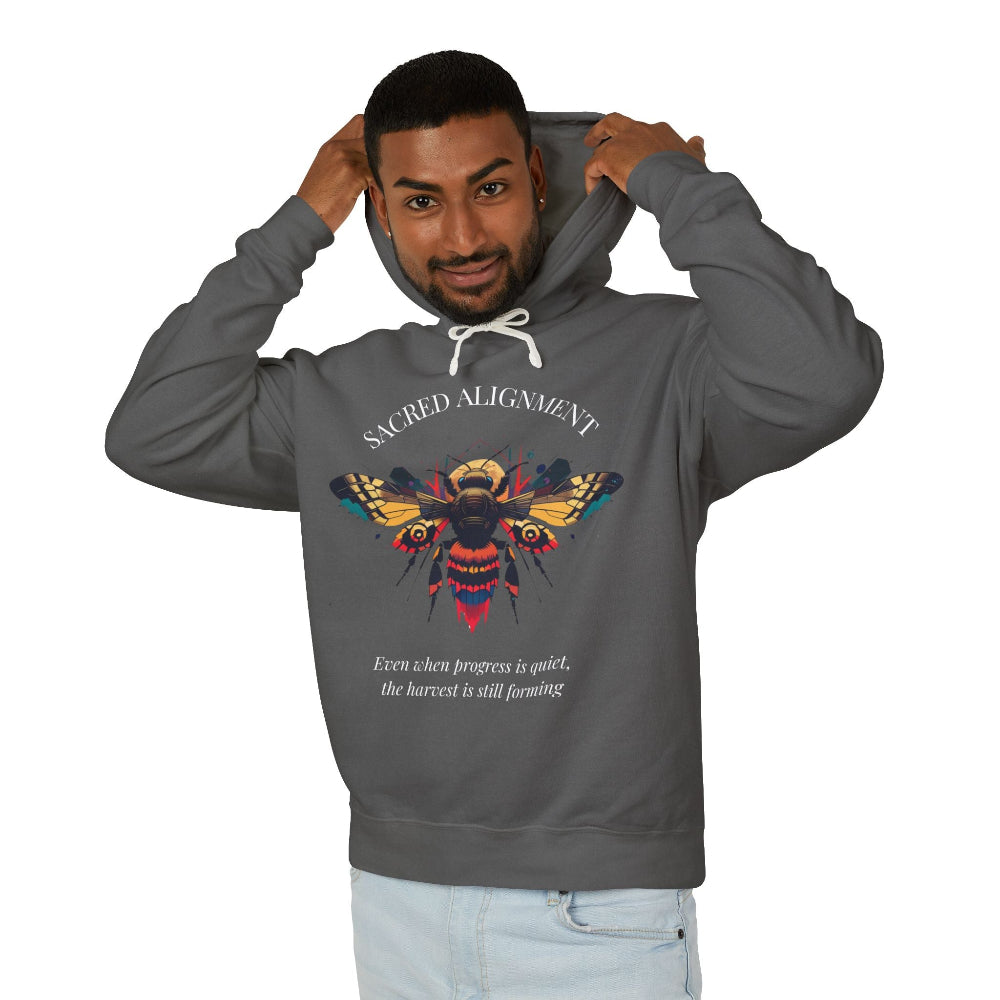 Man wearing a gray hoodie featuring a vibrant bee graphic with the text "SACRED ALIGNMENT" above and the quote "Even when progress is quiet, the harvest is still forming" below. Casual streetwear style with light jeans.