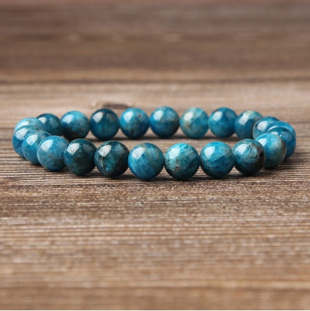 
Blue apatite gemstone bracelet: stack of polished round beads in vibrant turquoise-blue with natural inclusions, subtle chatoyancy, shiny faceted surfaces, displayed on a wooden background. Natural healing crystal jewelry for throat chakra, communication, and emotional clarity.