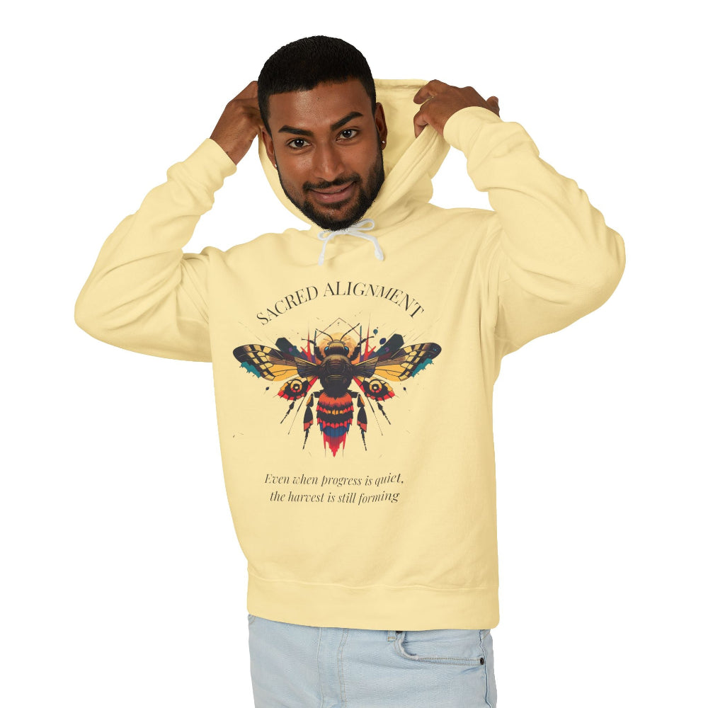 Man wearing a yellow hoodie featuring a vibrant bee graphic with the text "SACRED ALIGNMENT" above and the quote "Even when progress is quiet, the harvest is still forming" below. Casual streetwear style with light jeans.