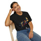 Black unisex cotton t-shirt modeled by smiling woman with braided hair, seated pose, hand on cheek, featuring bold Pisces zodiac graphic on chest: vibrant abstract twin goldfish swimming in mirrored formation, detailed scales and flowing fins, intense eyes, geometric accents, and dynamic multicolored paint splatters in gold, red, navy, teal, blue, and black tones bursting outward. Elegant black cursive "Pisces" text below. Astrology zodiac motivational crewneck tee.