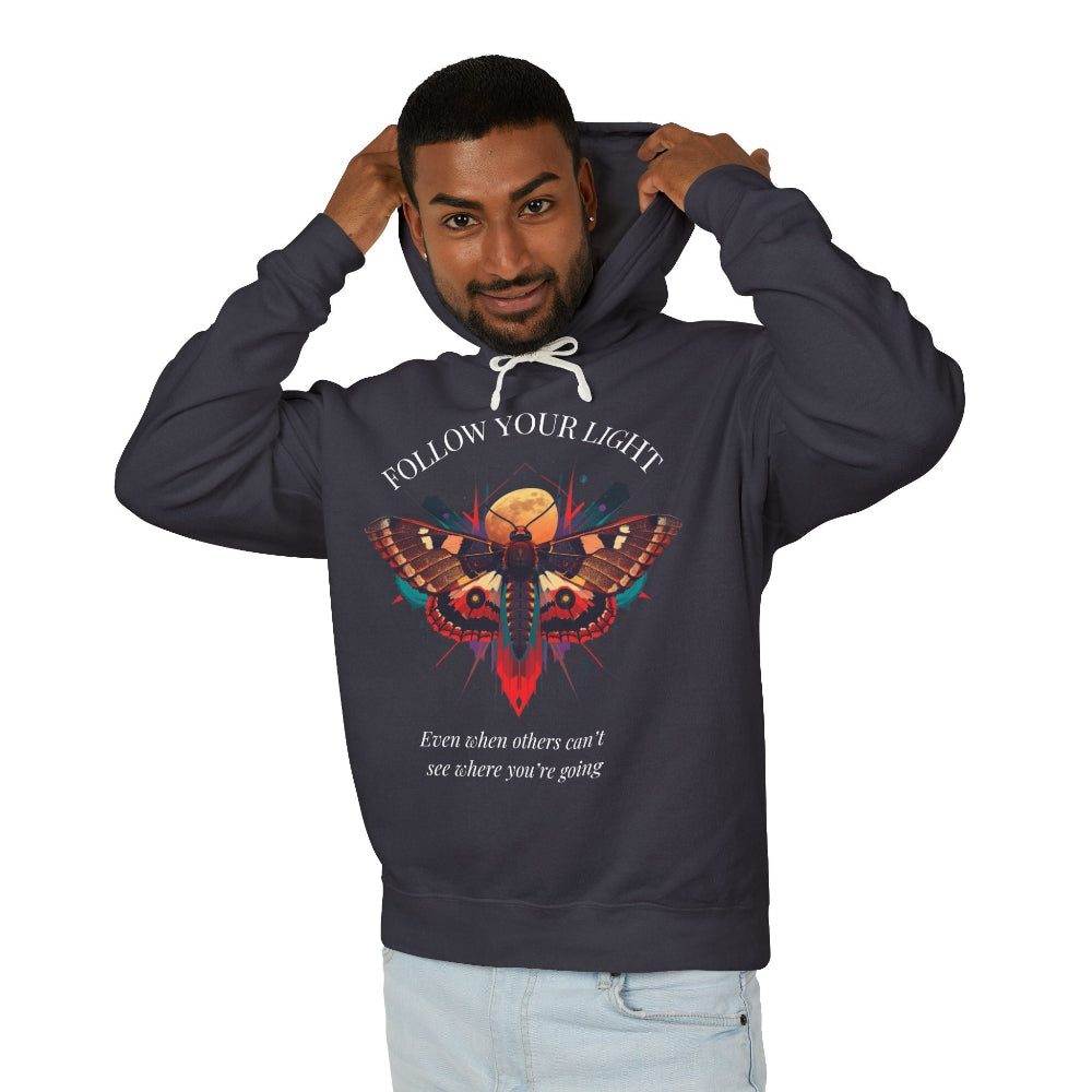 Young man with short hair pulling up the hood of a dark gray hoodie featuring vibrant multicolored moth/butterfly graphic, geometric sun/moon elements, "FOLLOW YOUR LIGHT" arched text above, motivational quote "Even when others can't see where you're going" below