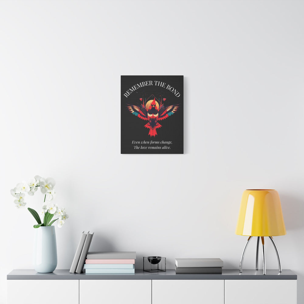 Minimalist interior scene featuring a framed wall art print with a colorful geometric phoenix and the quote "Remember the Bond – Even when forms change, the love remains alive." Styled on a light wall above a shelf with books and a yellow lamp.