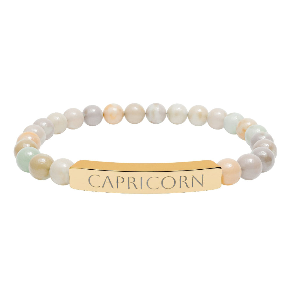 Bracelet with polished pastel beads in soft peach, cream, gray, and light blue tones with natural marbling, featuring a gold-plated curved bar engraved "CAPRICORN" in uppercase letters. Stretch-fit zodiac-inspired crystal jewelry.