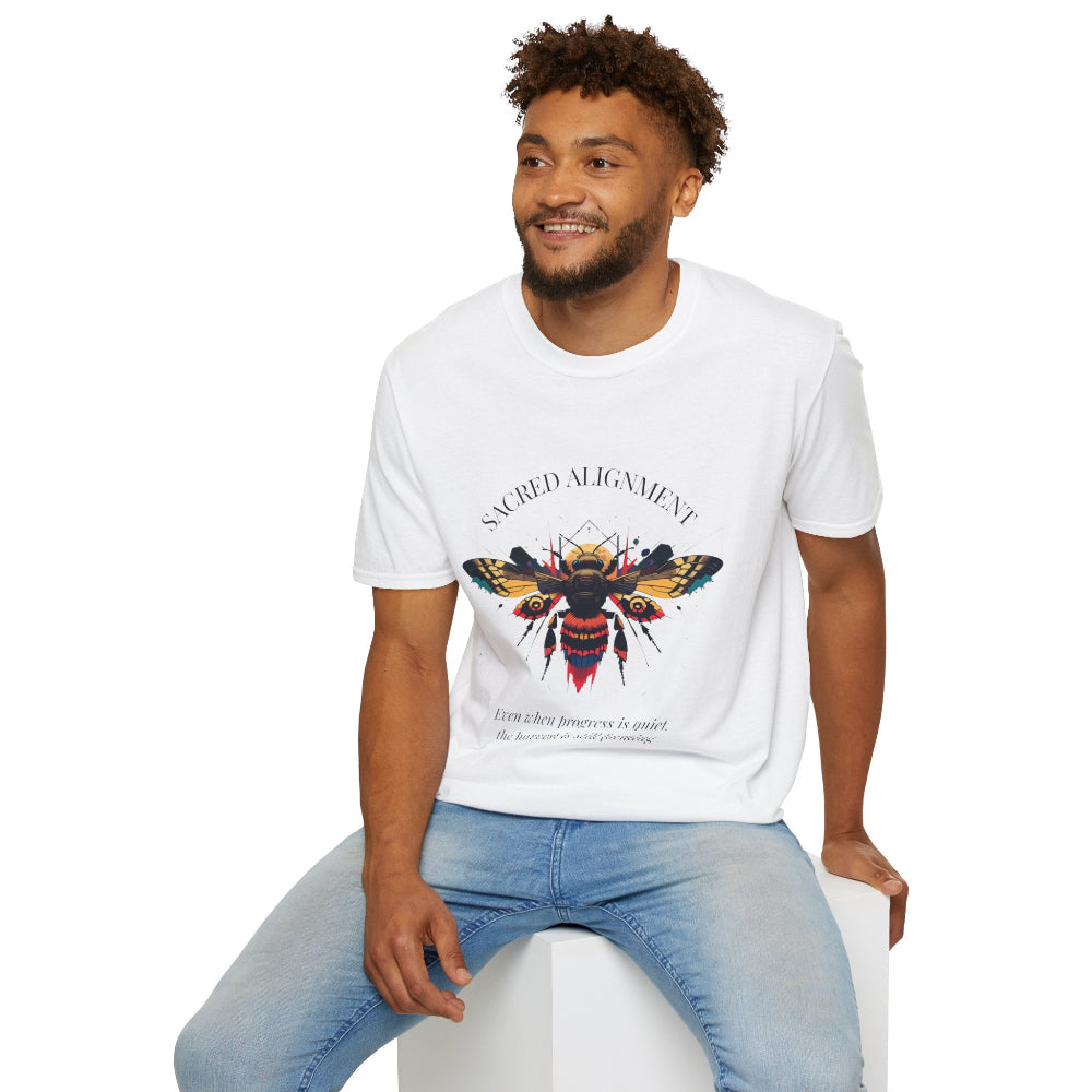 White unisex cotton t-shirt modeled by smiling man with curly hair and beard, featuring bold "Sacred Alignment" graphic on chest: vibrant abstract bee with detailed wings and body, colorful geometric accents, and dynamic paint splatters in red, orange, teal, blue, and gold tones around an orange moon. Arched white text "SACRED ALIGNMENT" above and smaller quote "Even when progress is quiet, the universe is conspiring" below. Motivational spiritual empowerment crewneck tee.