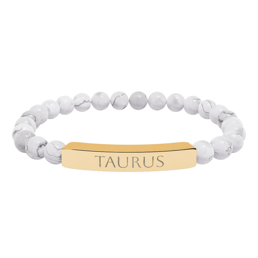 Bracelet with polished white howlite beads featuring natural gray-black veining and marbling, paired with a gold-plated curved bar engraved "TAURUS" in uppercase letters. Stretch-fit zodiac-inspired crystal jewelry.