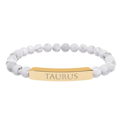 Bracelet with polished white howlite beads featuring natural gray-black veining and marbling, paired with a gold-plated curved bar engraved "TAURUS" in uppercase letters. Stretch-fit zodiac-inspired crystal jewelry.