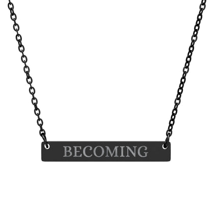 Black bar necklace with engraved uppercase text "BECOMING" on a matte black pendant, suspended on a delicate black chain. Minimalist motivational affirmation jewelry for growth, transformation, and self-evolution.
