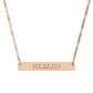 Rose gold bar necklace with engraved uppercase text "HEALED" on a sleek matte pendant, suspended on a delicate matching rose gold chain. Minimalist motivational affirmation jewelry for healing, recovery, and emotional restoration.