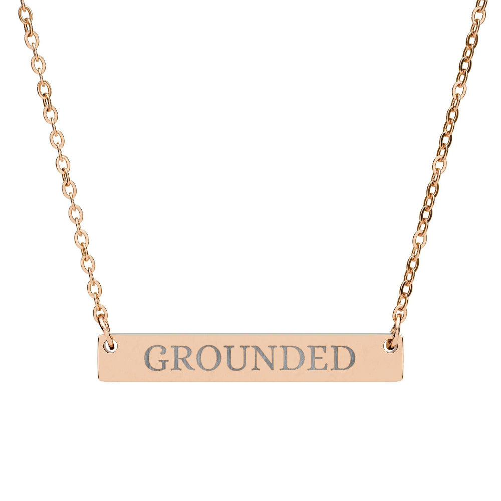 Rose gold bar necklace with engraved uppercase text "GROUNDED" on a sleek matte pendant, suspended on a delicate matching rose gold chain. Minimalist motivational affirmation jewelry for grounding, stability, and root chakra energy.