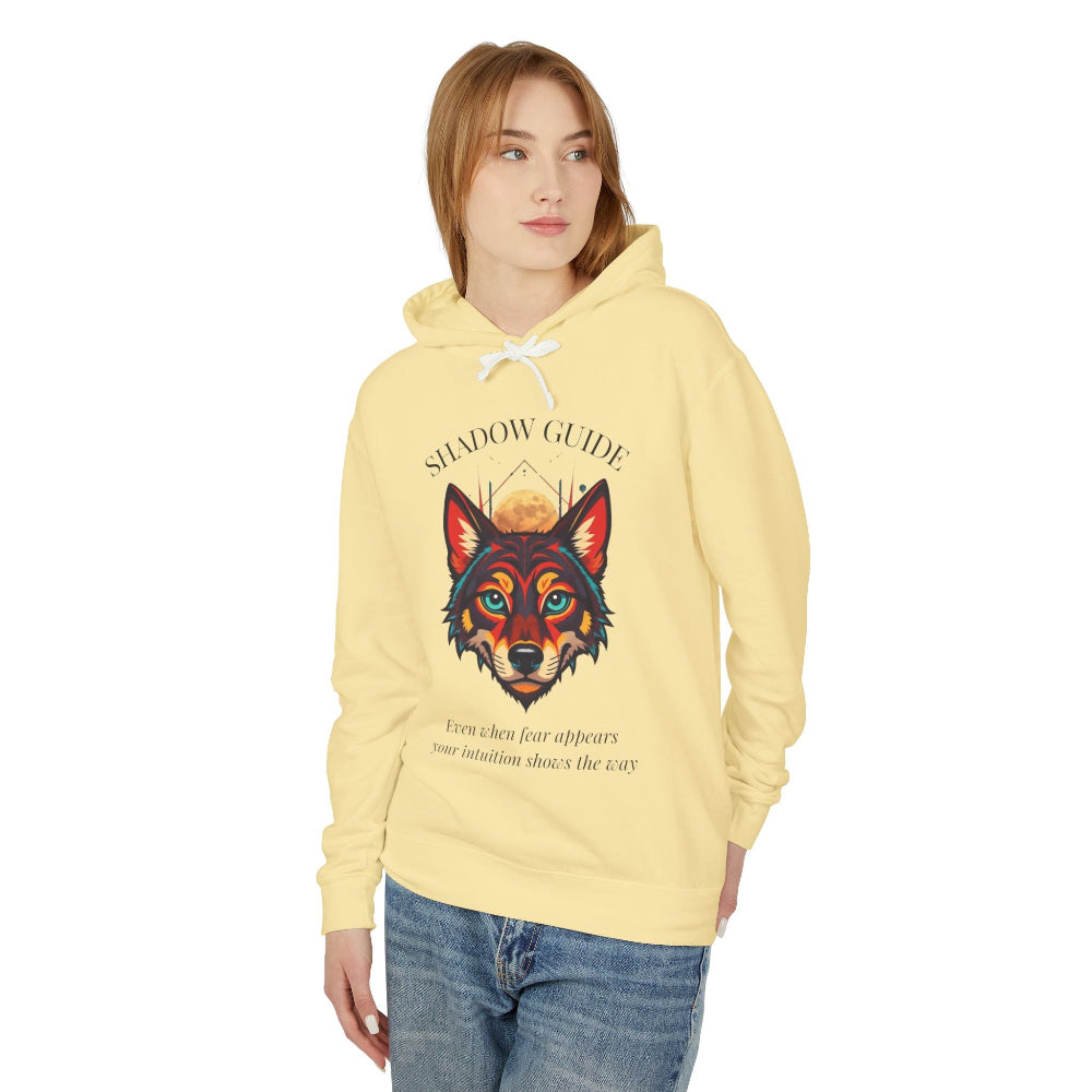 Young woman with long hair modeling pale yellow hoodie featuring vibrant multicolored wolf head graphic, intense eyes in orange blue teal splashes, full moon above, "SHADOW GUIDE" arched text, motivational quote about intuition and fear below
