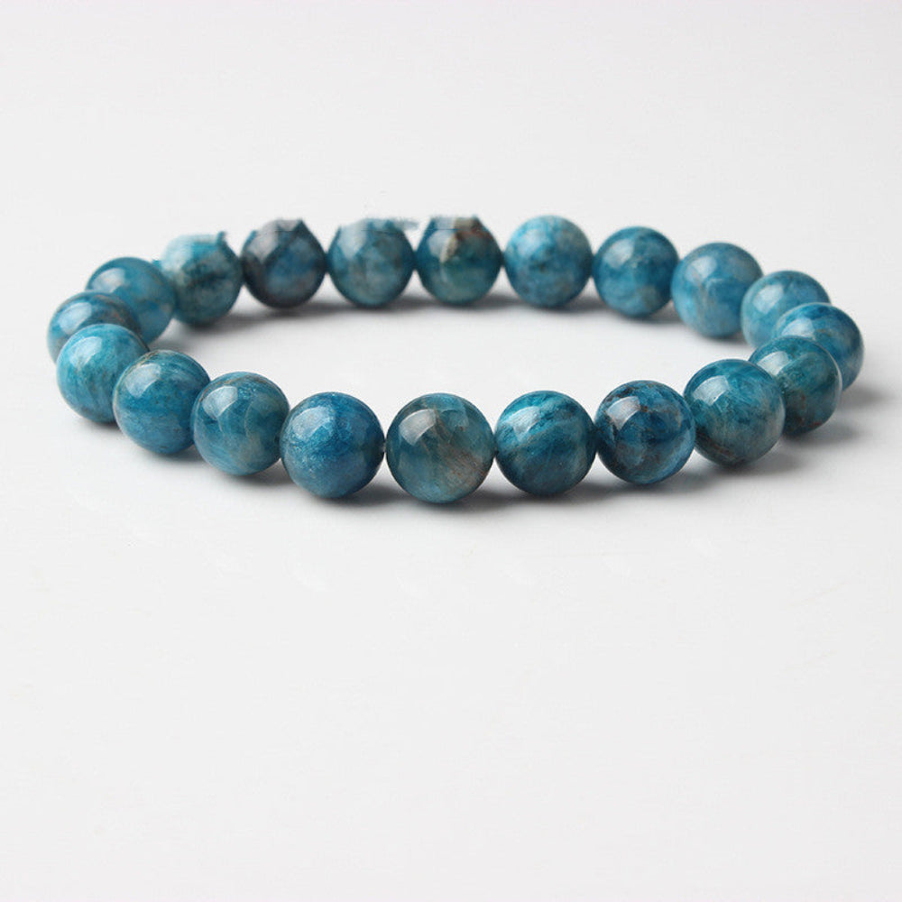 
Blue apatite gemstone bracelet: single strand of polished round beads in vibrant turquoise-blue with natural cloudy inclusions, faceted shiny surfaces, and subtle chatoyancy, displayed on a light wooden background. Natural healing crystal jewelry for throat chakra, communication, and emotional clarity.