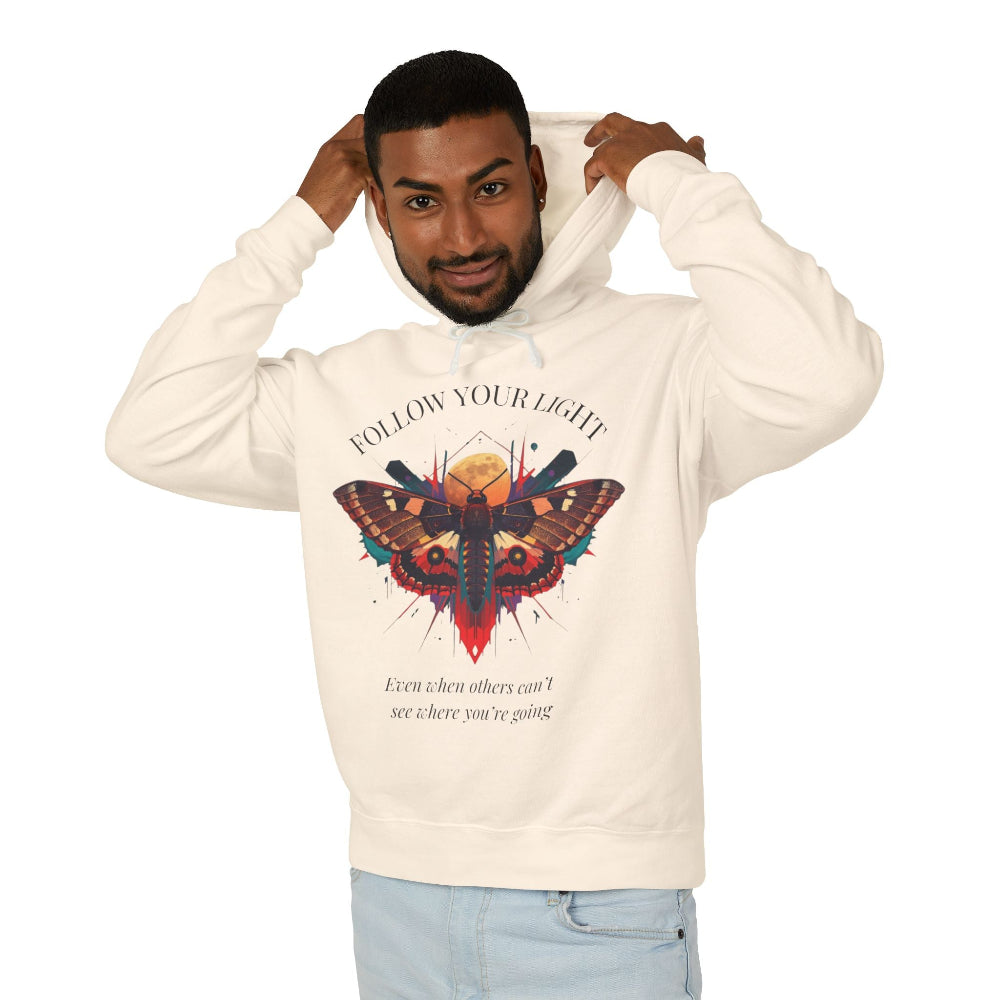 Young man with short hair pulling up the hood of a cream hoodie featuring vibrant multicolored moth/butterfly graphic, geometric sun/moon elements, "FOLLOW YOUR LIGHT" arched text above, motivational quote "Even when others can't see where you're going" below