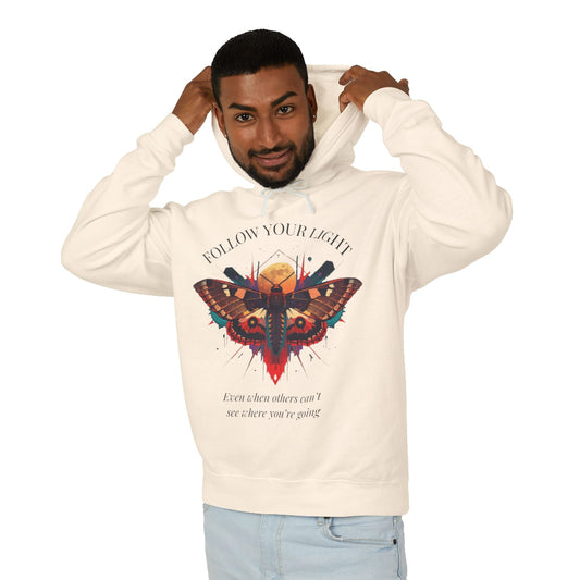 Young man with short hair pulling up the hood of a cream hoodie featuring vibrant multicolored moth/butterfly graphic, geometric sun/moon elements, "FOLLOW YOUR LIGHT" arched text above, motivational quote "Even when others can't see where you're going" below