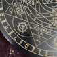 Close-up detail of a black and gold astrology wheel showing "Thursday - Tin - 2" with the Jupiter symbol, surrounded by directional markers, planetary associations, and intricate sacred geometry designs. Pagan divination board with metallic gold engraving.