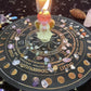 Angel-shaped candle with a lit flame placed in the center of a black and gold Wheel of the Year board, surrounded by scattered crystal chips and tumbled stones. Mystical pagan altar setup with sabbat names and zodiac symbols. Spiritual seasonal decor.