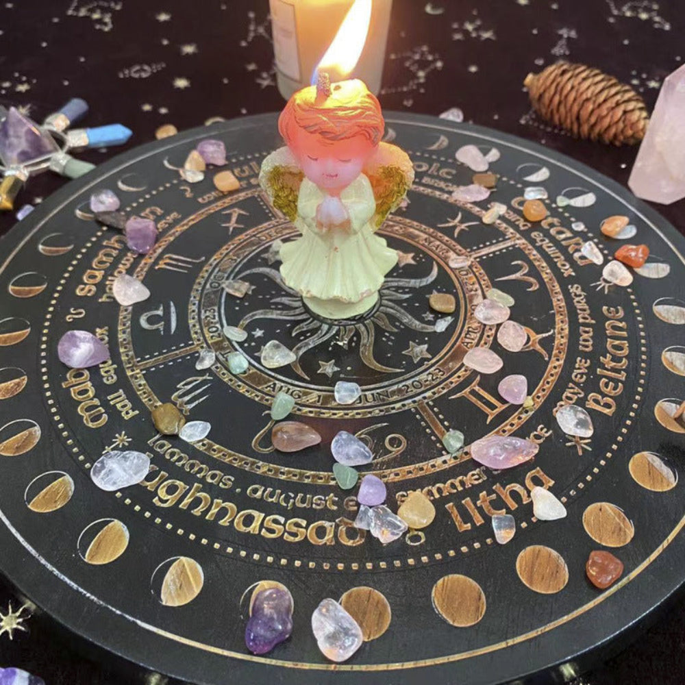 Angel-shaped candle with a lit flame placed in the center of a black and gold Wheel of the Year board, surrounded by scattered crystal chips and tumbled stones. Mystical pagan altar setup with sabbat names and zodiac symbols. Spiritual seasonal decor.