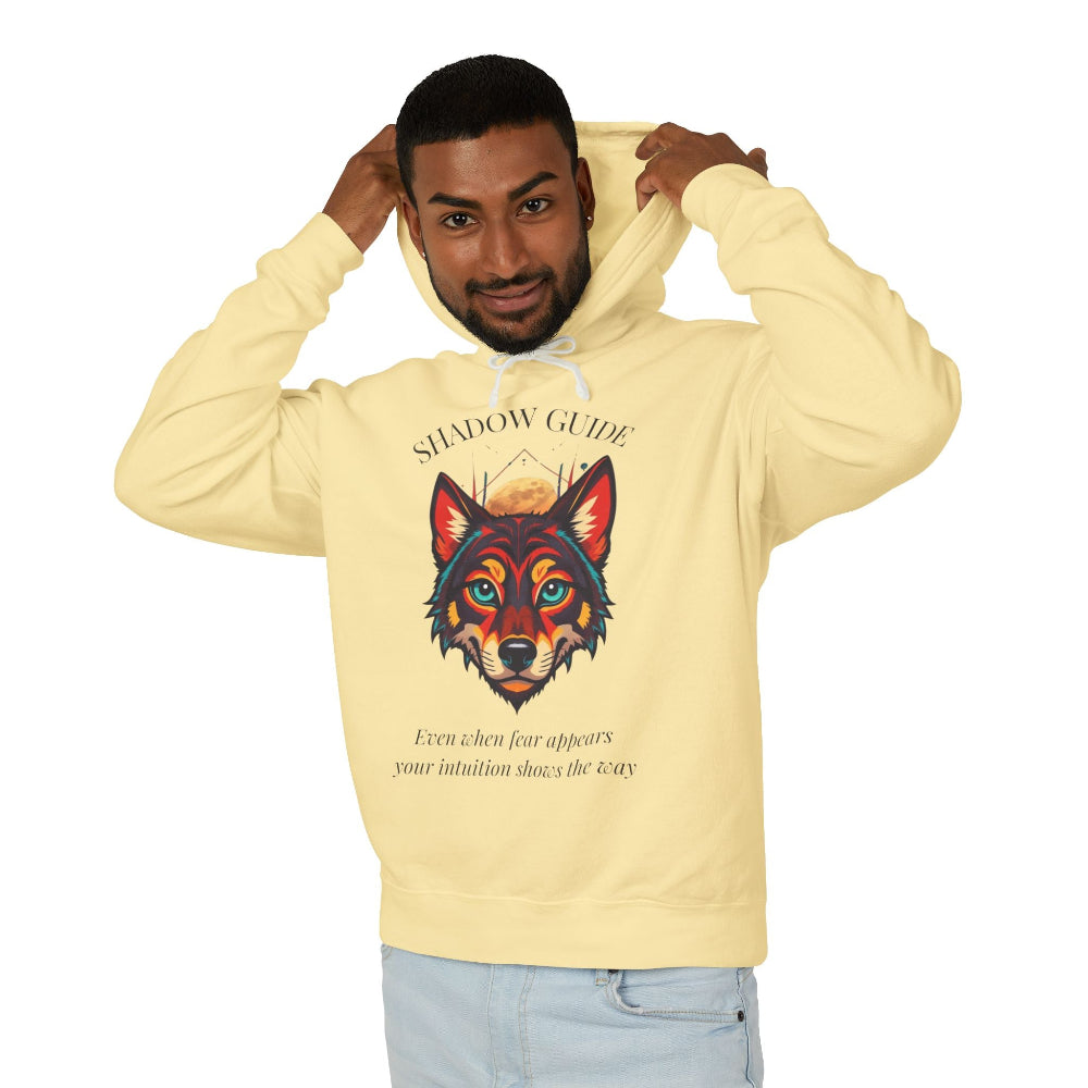 Young man with short hair pulling up the hood of a pale yellow hoodie featuring vibrant multicolored wolf head graphic, intense eyes in orange blue teal splashes, full moon above, "SHADOW GUIDE" arched text, motivational quote about intuition and fear below