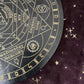 Close-up detail of a black and gold astrology wheel board showing planetary days (Tuesday - Iron, Wednesday - Mercury), directional elements (East - Air), and sacred symbols including the Om symbol and planetary glyphs. Intricate pagan divination tool on a dark starry background.