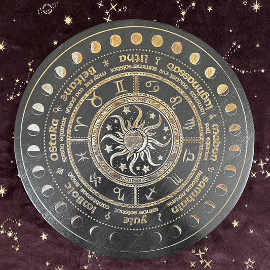 Black and gold Wheel of the Year divination board featuring a central sun and moon design surrounded by zodiac symbols, sabbat names (Yule, Imbolc, Ostara, Beltane, Litha, Lammas, Mabon, Samhain), and moon phase markers. Pagan seasonal wheel on a dark velvet background with gold stars.